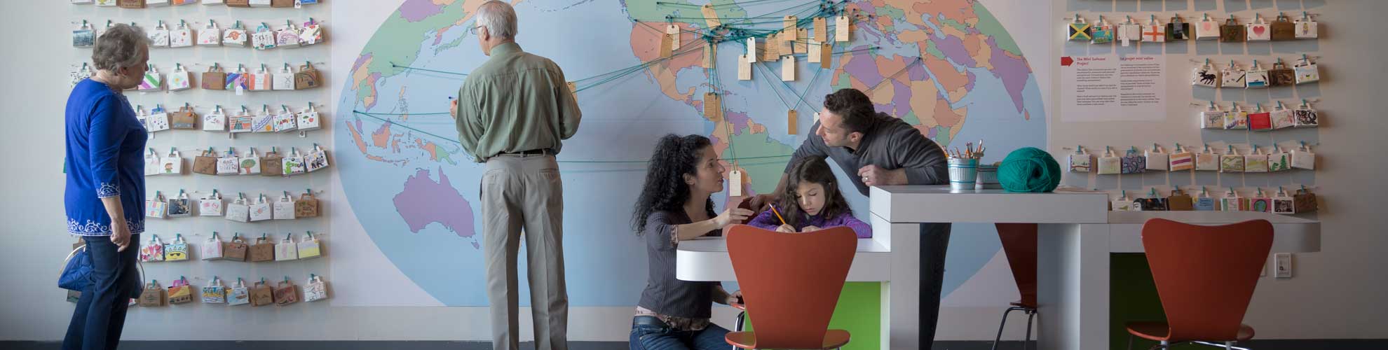 Five adults and one child interact with the exhibition, featuring a map of the world with luggage tags.