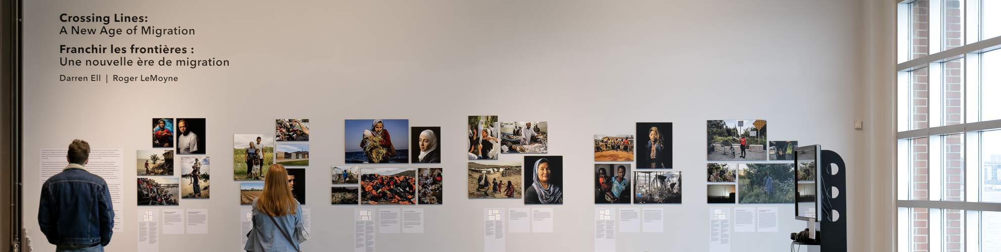 Two people look at a wall of photographs and text for the Crossing Lines exhibition.