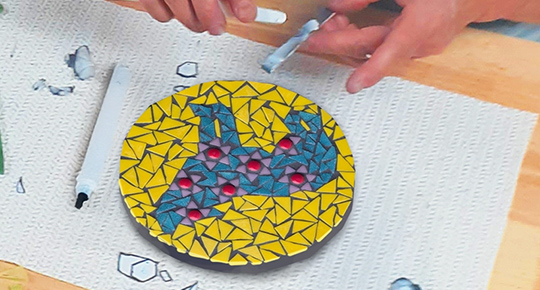 A participant carefully places coloured glass tiles onto a circular mosaic panel featuring a teal wolf design on a yellow background.