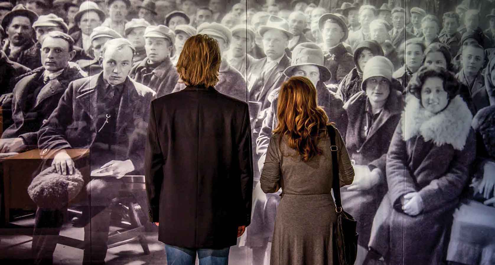Man and woman stare at a life-size historical photograph of immigrants waiting to be called.