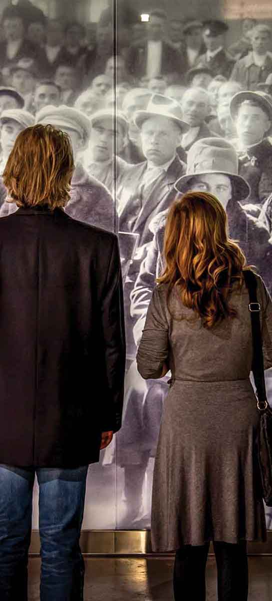 Man and woman stare at a life-size historical photograph of immigrants waiting to be called.