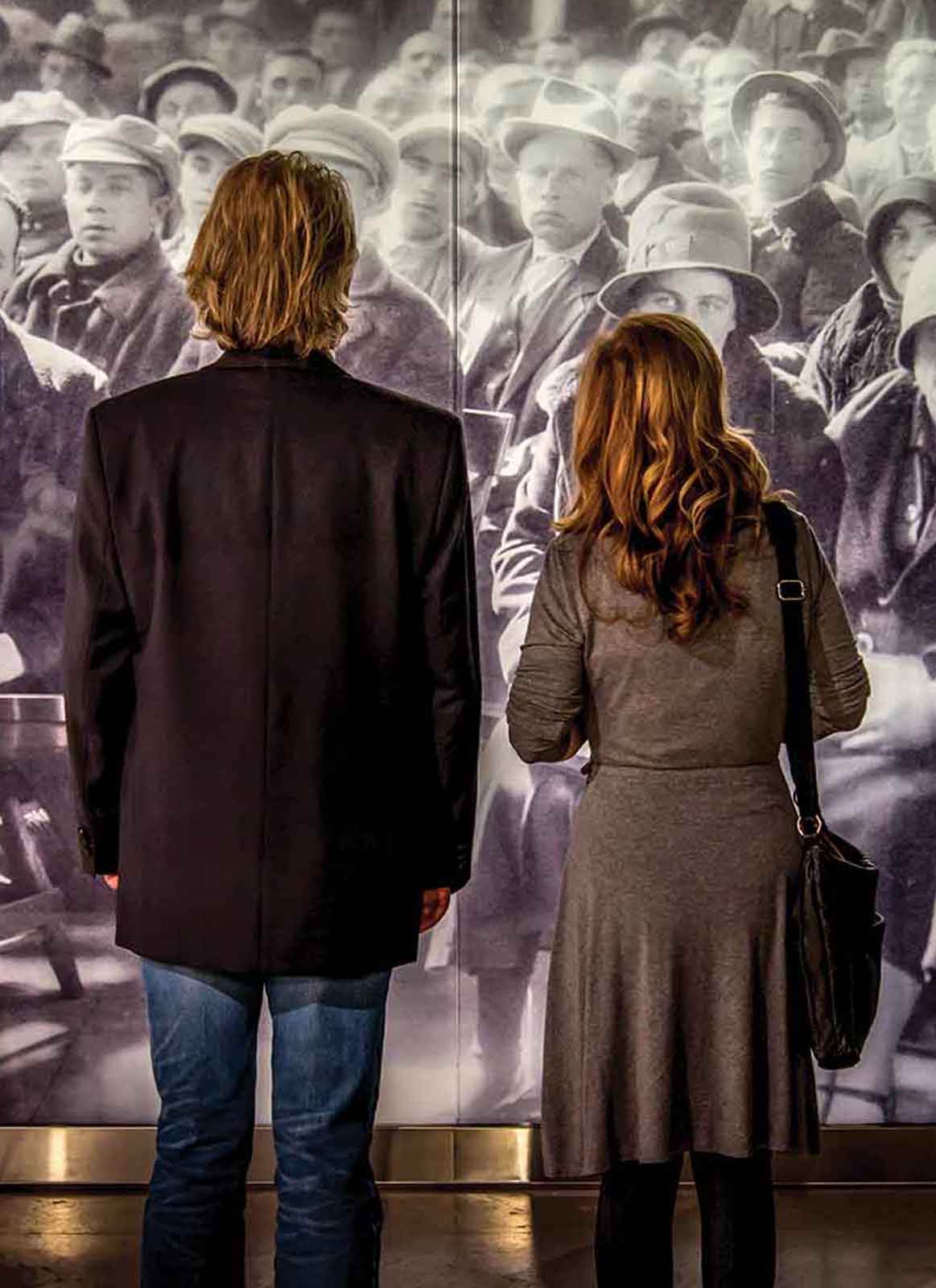 Man and woman stare at a life-size historical photograph of immigrants waiting to be called.