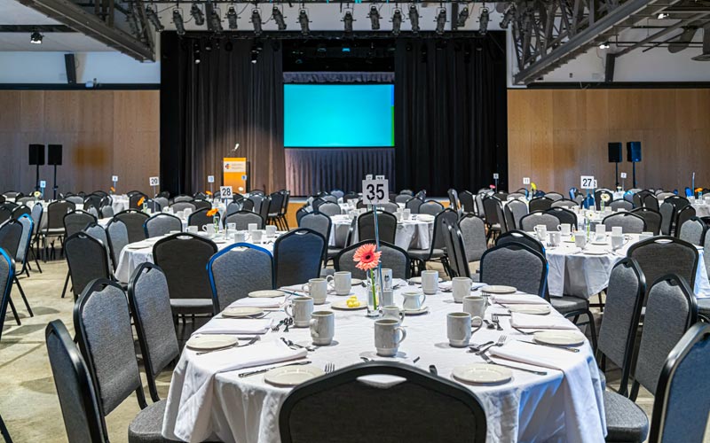 The venue space is set for a banquet dinner with oval tables and banquet chairs. Each table has a table number sign and a bud vase with a daisy. There is a stage with a podium with microphone, and a projection screen.