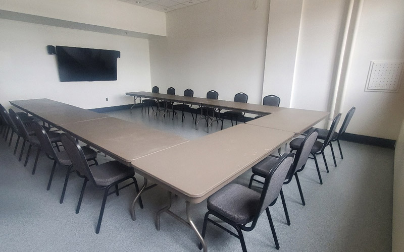 A small room with tall ceilings set with rectangular tables and banquet chairs in a U-shape for fifteen guests.  The set-up is facing a TV for a presentation.