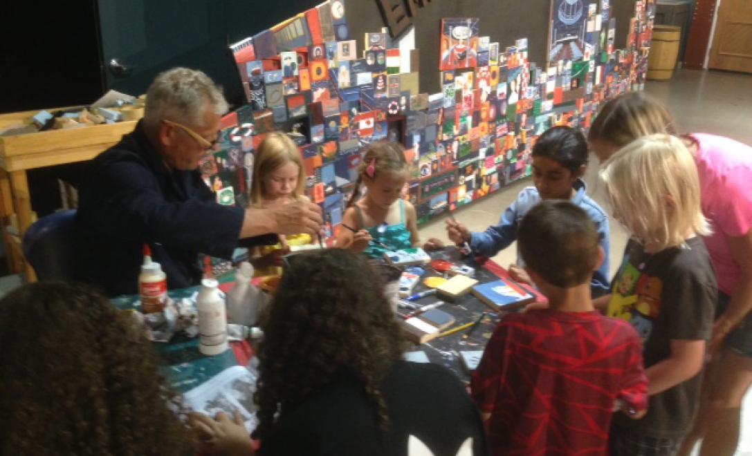 A man sits at a table with a group of children and is showing them how to paint.