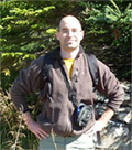 Man, dressed in khaki shirt and pants carrying a backpack, stands on rocky terrain