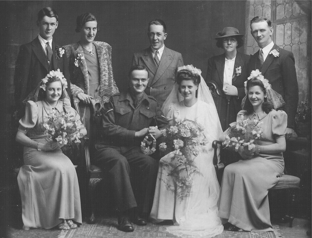 Archived portrait of three smiling brides sitting with bouquets, their grooms stand behind.