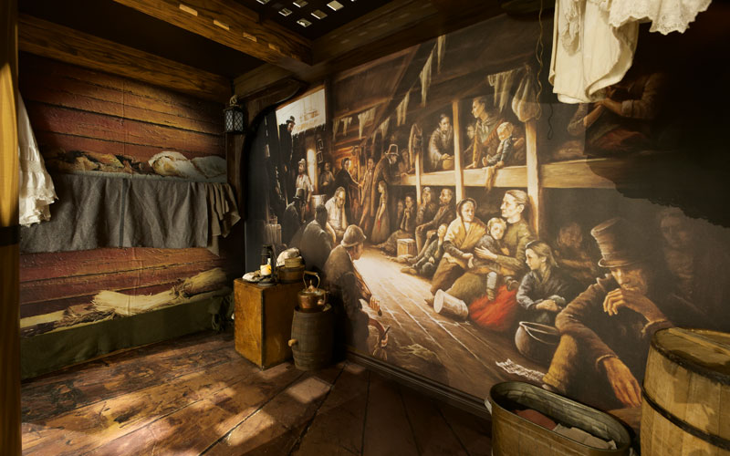 A model room showing the inside of an old ship, with several props and a large picture of people in bunks.