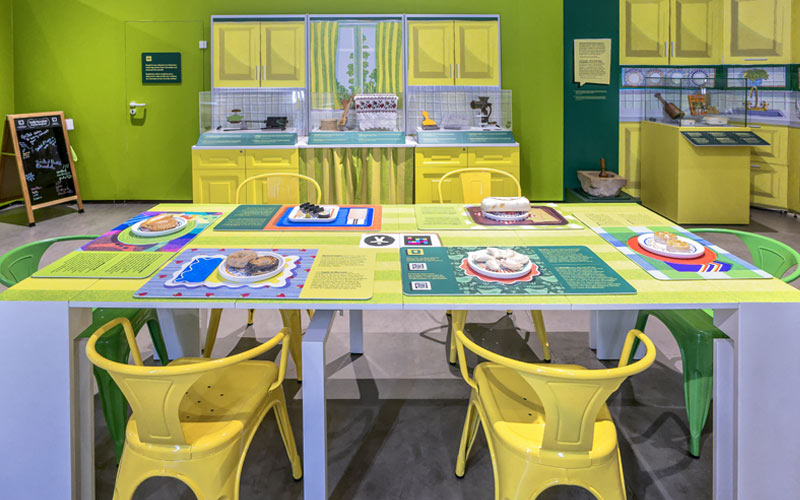 A green kitchen featuring a table with model foods on plates, including bannock, sushi, and perogies.