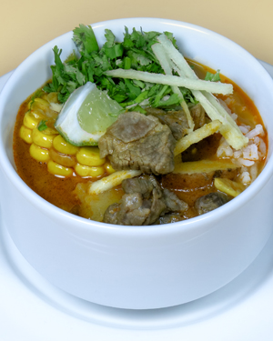 A bowl of aloo birria with reddish broth, corn on the cob, beef pieces, rice, cilantro, tortilla strips and a lime wedge.