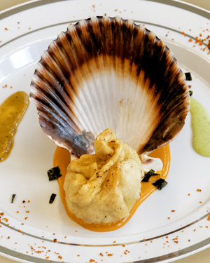 A seafood momo placed in front of a scallop shell on a white plate, set on an orange sauce base, with garlic cilantro sauce on the left and timur chutney sauce on the right.