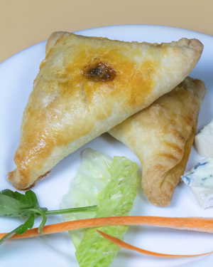 A white plate holding two golden-brown Ukajas alongside thin carrot sticks, a lettuce leaf, a sprig of parsley and two wedges of white cheese.