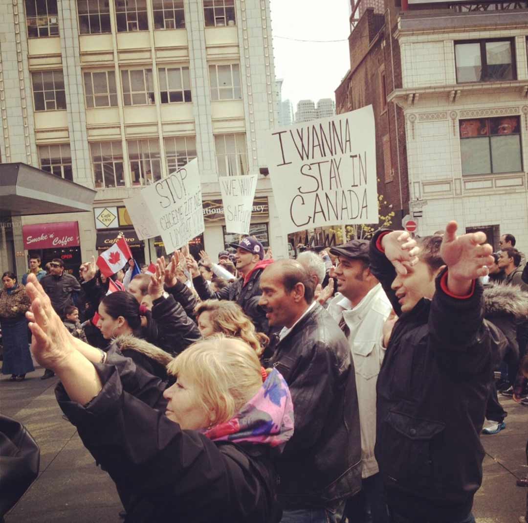A crowd of people walk through a city street holding protest signs, one sign reads I wanna stay in Canada.