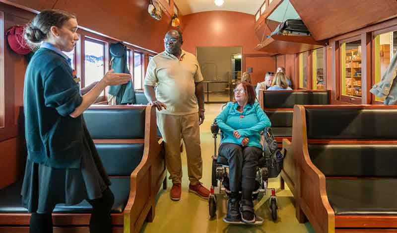 A museum guide speaks with two visitors, one in a wheelchair, inside a historic train car exhibit with wooden benches.