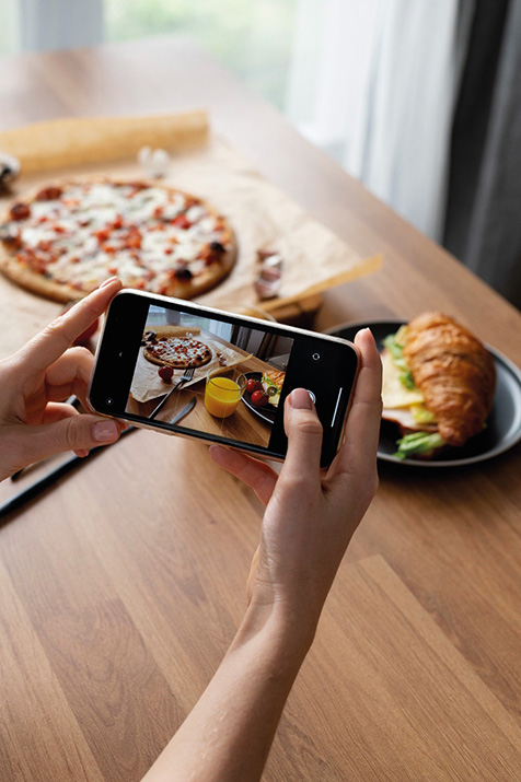 A person holds a smartphone and takes a photo of a meal on a wooden table, including a pizza, a croissant sandwich on a plate, a glass of orange juice, and some cherry tomatoes.