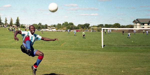 A black man smiles mid-air after kicking a ball, in bright sun on a large field with an active soccer pitch behind him.