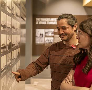 A man and woman point to a ship image on a wall of ship photos.