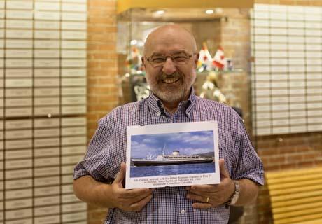 An older man holds up a ship photo in front of the Wall of Honour plaques.