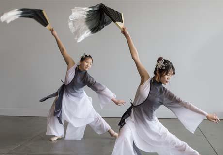 Two Asian dancers in flowing costumes wave the same flowing material above their heads as they move.
