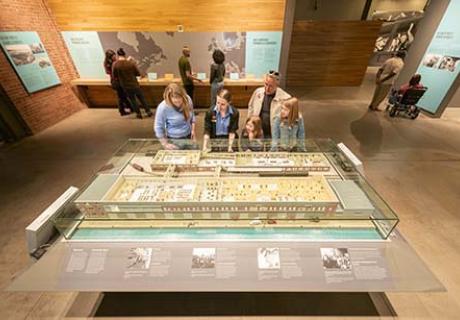 An interpreter, a man and a woman are discussing the table-sized model of historic Pier 21.