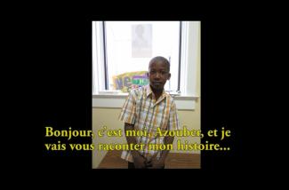 A young boy in a checkered shirt stands in front of a window.