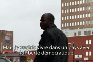 A young man stands in front of a building and is looking over his shoulder.