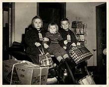 Black and white image of three kids sitting with luggage bags, they are holding each other and look very happy.