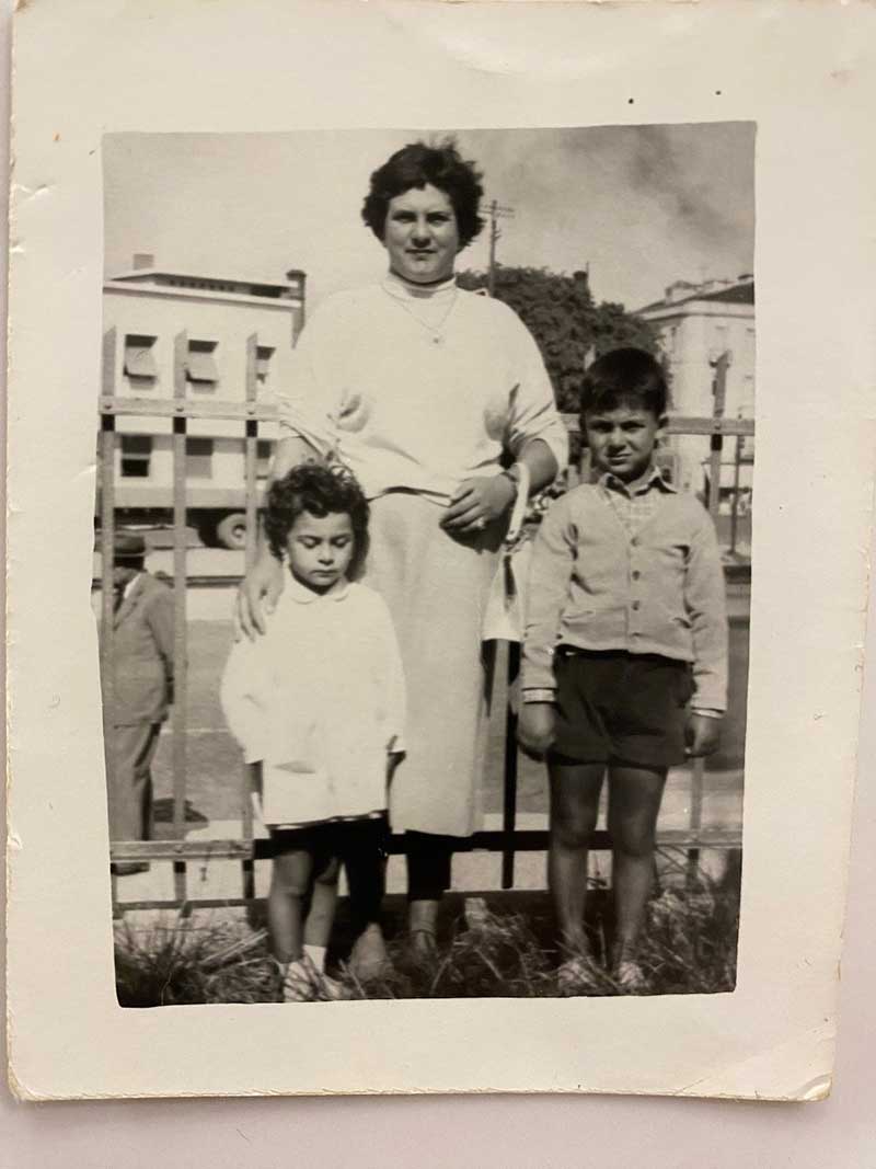 A woman and two children pose for the camera, buildings can be seen in the background.