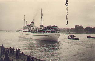 Ship departing dock, smaller boats alongside and people watching from dockside.