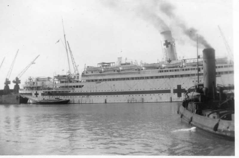 Ship with a red cross sign is docked and there is a small ship in front of it.