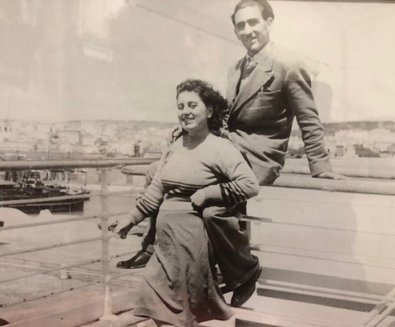 A young man sits on the railing of a ship's deck and a young woman stands in front of him.