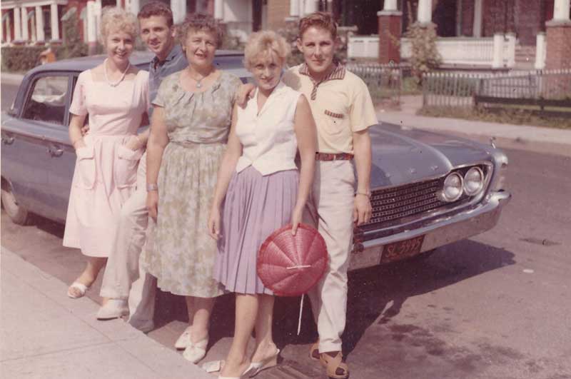 Three women and two men lean against an old blue car.