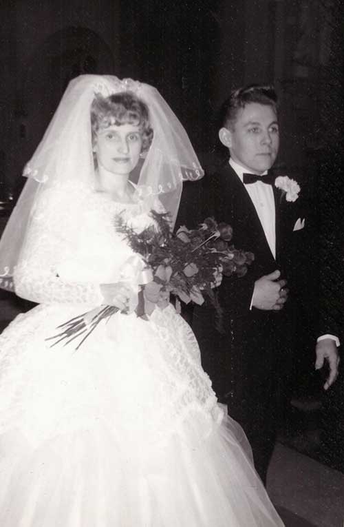 A bride and groom on their wedding day, the bride carries a bouquet and wears a veil.