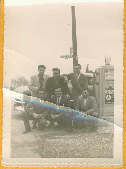 Six young men sitting and standing next to a gas pump.