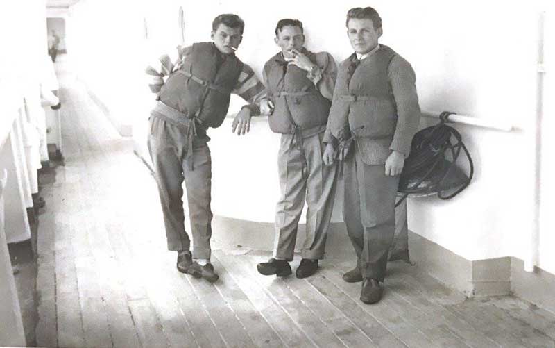 Three men in life vests smoke cigarettes on the deck of a ship.