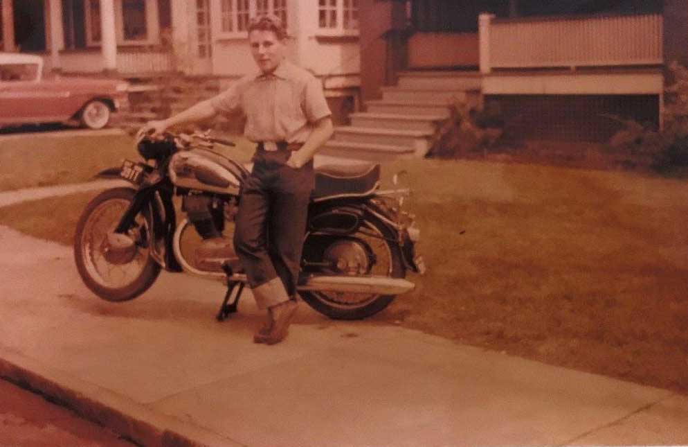 A young man leans against a motorcycle with one hand on the steering and one hand in his pocket.