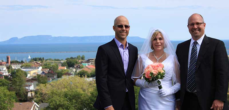 A young bride is standing between two well-dressed men; she is wearing a veil and dress and holds a bouquet of coral-coloured roses.
