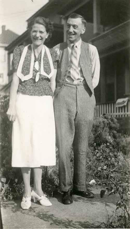 A smiling couple stand in front of the hedge of their house.