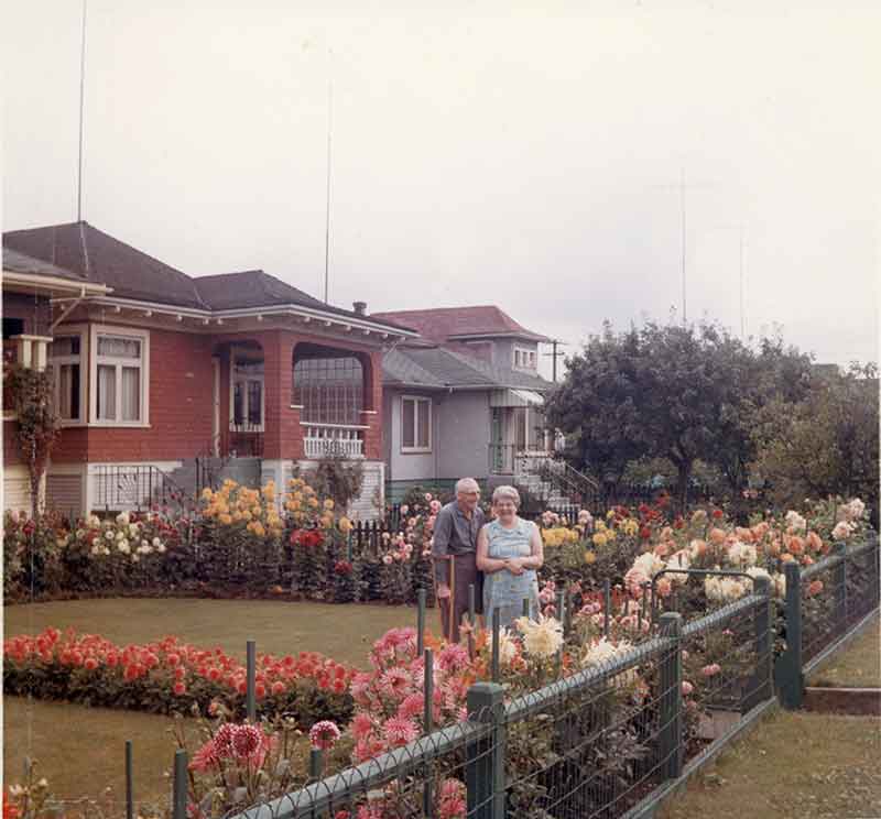 A man and woman stand at the bottom of their garden, that is filled with beautiful flowers.