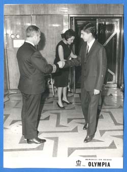 A man in black suit shaking the hand of captain of ship.