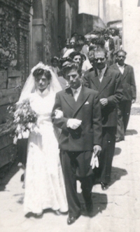 Bridal procession walking through a narrow street with bride and groom leading.