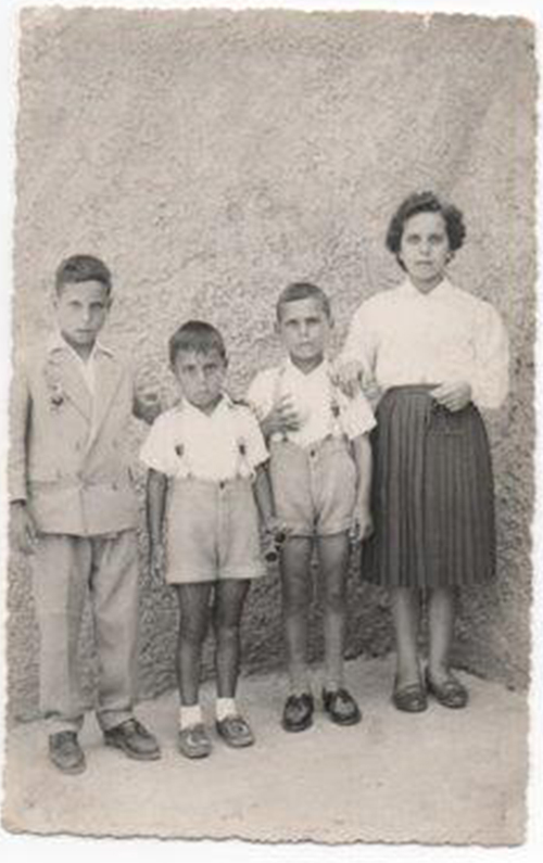 Old black & white photo of three kids standing with young lady in front of wall for photo.