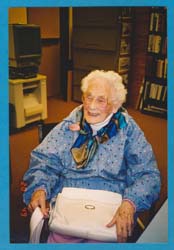 Grace seated, wearing bright blue jacket and holding big white purse.