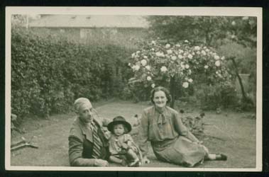 Young Joan wearing a hat, sitting in the grass of a garden with her parents.