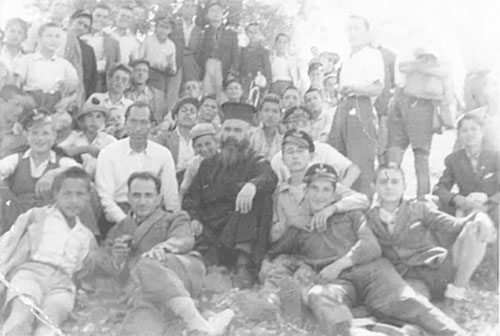 A large group of people are sitting on a hall, in the center there is a man dressed in black religious clothing.