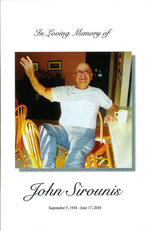 An elderly man sits on a wooden chair and waves happily at the camera.
