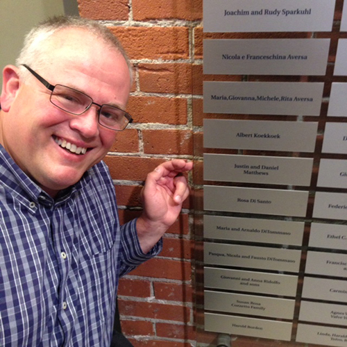 A handsome smiling man is kneeling close to the Wall of Honour at Pier 21 Museum.