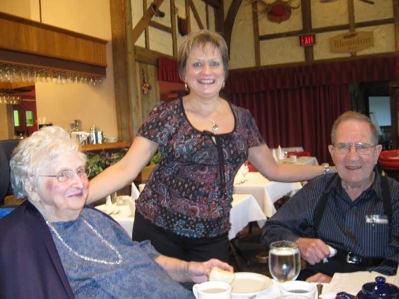A couple are seated at a table and a woman stands between them with a hand on both of their shoulders.