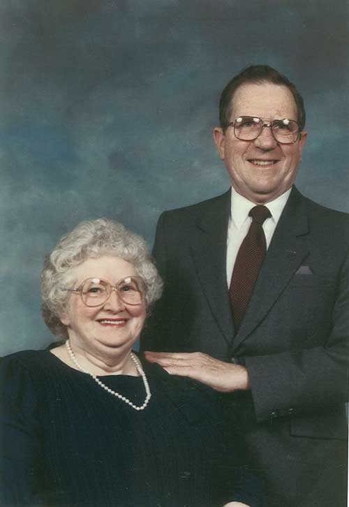 Coloured portrait of a couple, the woman is seated and wears a pearl necklace and the man stands next to her.