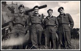 Four young men in military uniform and beret, standing in front of an army tank.
